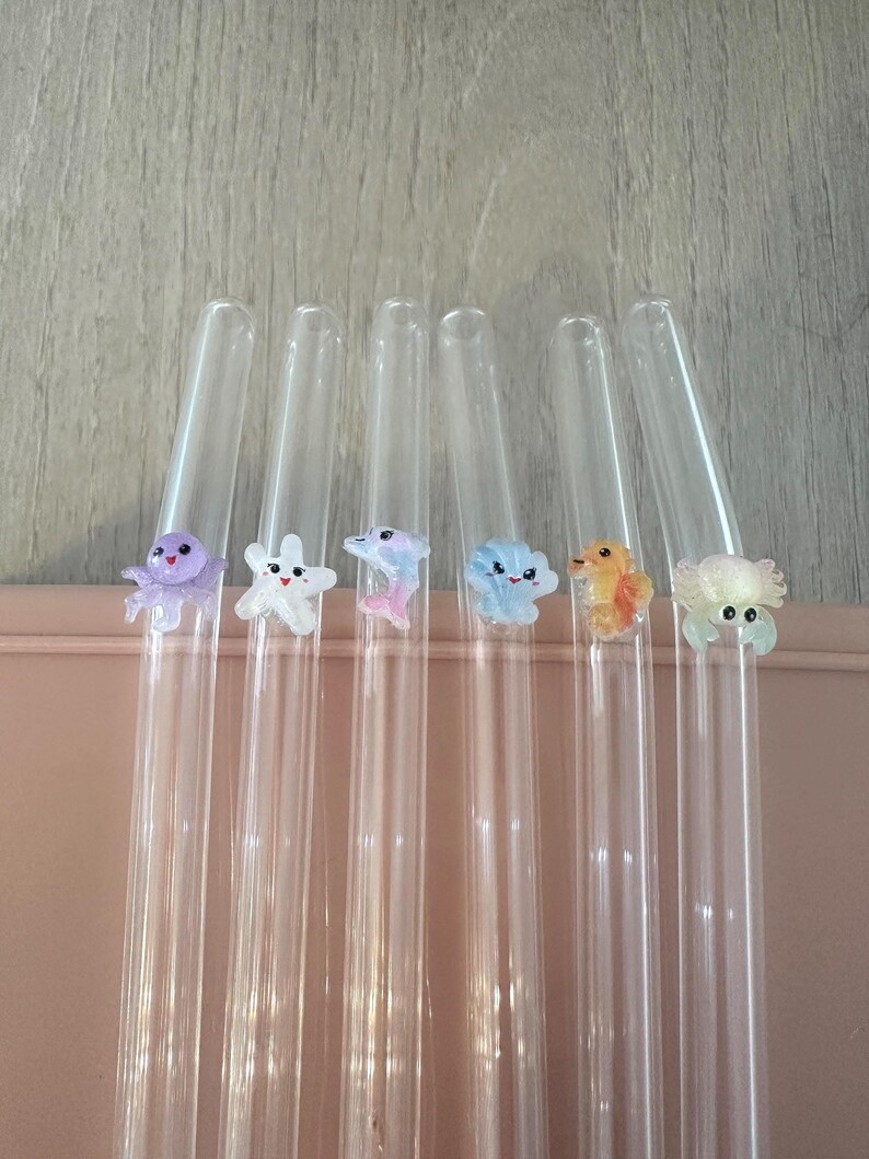 May include: Six clear glass drinking straws, each featuring a small, colorful, three-dimensional sea creature figurine attached to the side. The figures include a purple octopus, a white starfish, a pink dolphin, a blue shell, an orange seahorse, and a crab.
