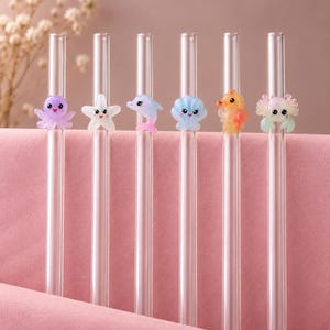 May include: Six clear glass drinking straws, each featuring a small, colorful, cartoon-style sea creature charm. The charms include a purple octopus, a white starfish, a pink and blue dolphin, a blue seashell, an orange seahorse, and a crab.
