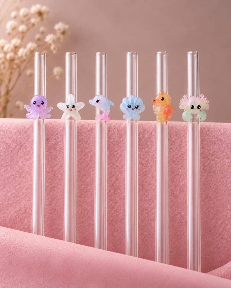 May include: Six clear glass drinking straws, each featuring a small, colorful, cartoon-style sea creature charm. The charms include a purple octopus, a white starfish, a pink and blue dolphin, a blue seashell, an orange seahorse, and a crab.