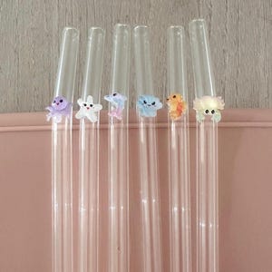 May include: Six clear glass drinking straws, each featuring a small, colorful, three-dimensional sea creature figurine attached to the side. The figures include a purple octopus, a white starfish, a pink dolphin, a blue cloud, an orange fish, and a crab.