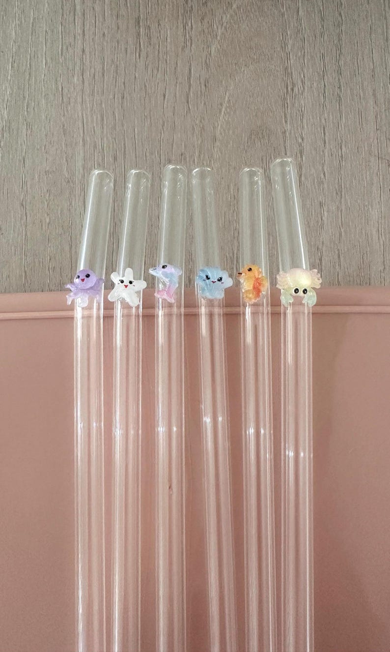 May include: Six clear glass drinking straws, each featuring a small, colorful, three-dimensional sea creature figurine attached to the side. The figures include a purple octopus, a white starfish, a pink dolphin, a blue cloud, an orange fish, and a crab.