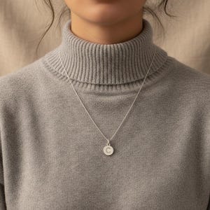 Puede incluir: Un collar de plata con un pequeño colgante redondo con la letra "C". El colgante cuelga de una fina cadena. La persona lleva un jersey de cuello alto gris.