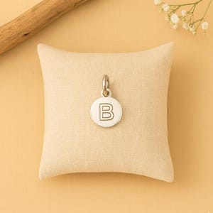 Pode incluir: Pingente prateado com a letra "B" gravada em um berloque redondo. O pingente pende de um pequeno laço e repousa sobre uma almofada bege. O fundo é laranja claro, com um pequeno ramo de flores brancas.