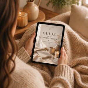 Peut inclure: Une personne tient une tablette affichant la couverture d'un livre avec le texte "GUIDE POUR LES CORPS FATIGUÉS". La tablette est tenue sur une couverture beige. Une bougie allumée, un vase avec des fleurs séchées et une tasse sont visibles.