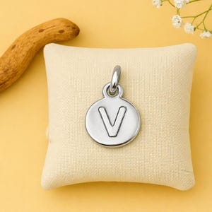Pode incluir: Pingente prateado com a letra "V" gravada em um berloque redondo. O pingente pende de uma pequena argola e é exibido em uma almofada bege. O fundo é amarelo suave.