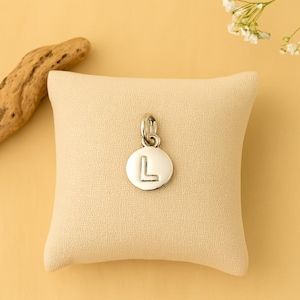 Puede incluir: Un charm de color plateado con la letra "L" grabada en un colgante redondo. El charm se muestra sobre un cojín beige, con un trozo de madera flotante y flores blancas en el fondo. El charm es probablemente para un collar o pulsera.