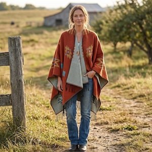 Peut inclure: Une femme porte un poncho rouille orné de motifs géométriques turquoise et dorés. Le poncho, avec une bordure à franges, est superposé à un haut gris clair et un jean bleu. L'arrière-plan montre une clôture en bois, une grange et un champ.