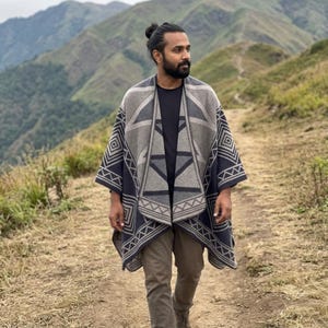 Peut inclure: Un poncho à motifs géométriques gris, noir et blanc. Le poncho est porté sur un t-shirt noir et un pantalon marron. L'arrière-plan présente une chaîne de montagnes et un sentier de terre.