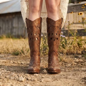 以下が含まれることがあります： 茶色の革のカウボーイブーツ。装飾的な切り抜きとスタッズが施されています。ブーツはポインテッドトゥで、膝丈です。背景には木の柵と乾燥した草が含まれています。