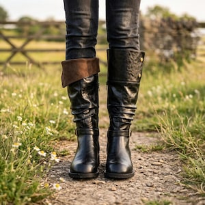 May include: Black leather knee-high boots with a brown suede cuff. The boots have a buckle detail at the ankle and gold-colored button accents. The boots are worn with dark denim jeans.