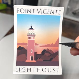 May include: A rectangular sticker featuring a lighthouse illustration. The design includes a pink lighthouse, brown cliffs, and a blue and orange sky. The text "POINT VICENTE LIGHTHOUSE" is printed on the sticker.