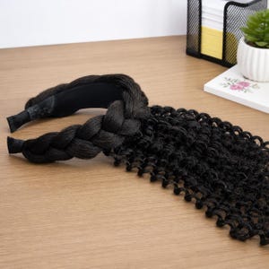 May include: A black braided headband with attached curly hair extensions. The headband is made of black fabric and features a thick, textured braid. The curly hair extensions are a dark black colour and have a natural, wavy appearance.