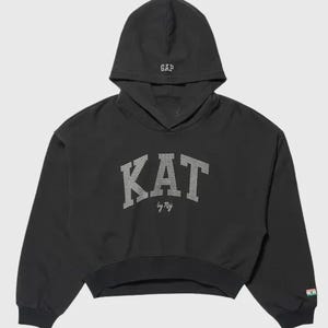 Puede incluir: Una sudadera negra corta con capucha que tiene el texto GAP bordado en blanco. El frente muestra la palabra KAT en pedrería plateada con el texto by Ref debajo. Un pequeño parche de bandera es visible en el puño de la manga derecha.