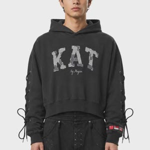 Puede incluir: Una sudadera con capucha gris carbón que presenta el texto KAT en una fuente gris desgastada en el pecho, con el texto en cursiva by Mayan debajo. Las mangas largas incluyen cordones negros con ojales metálicos y pequeños parches de banderas.