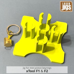May include: A bright yellow laser jig for mini piston keyrings, featuring a square base with multiple vertical semi-circular slots. A gold-coloured metal piston keyring is positioned to the left, with a measurement arrow indicating 11mm.