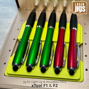 May include: Five pens with black grips and silver clips, featuring three green and two red barrels, are arranged in a bright yellow plastic jig. The jig is designed for xTool F1 and F2 machines, with the text Laser Jigs visible in the corner.