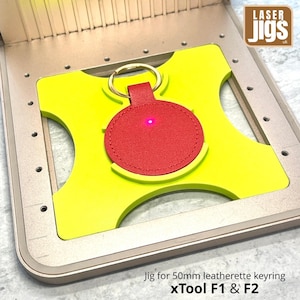 May include: A bright lime green jig for a 50mm leatherette keyring, featuring a circular cutout holding a red circular leatherette keyring with a gold metal ring. A small red laser dot is visible on the surface of the red keyring.