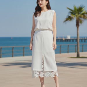 Crisp white Tavya high-waist midi skirt with an A-line silhouette and a front slit revealing intricate floral lace, worn by a model outdoors with an ocean view.