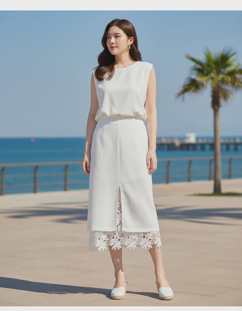 Crisp white Tavya high-waist midi skirt with an A-line silhouette and a front slit revealing intricate floral lace, worn by a model outdoors with an ocean view.
