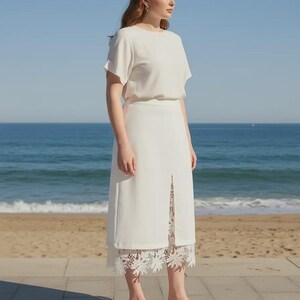 Crisp White Tavya high-waist midi skirt featuring a front slit and delicate floral lace underlay, modeled in a side view against a bright ocean background.