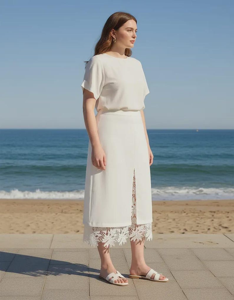 Crisp White Tavya high-waist midi skirt featuring a front slit and delicate floral lace underlay, modeled in a side view against a bright ocean background.