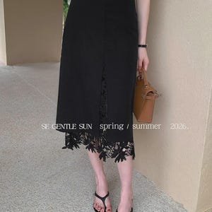 Model wearing a black Tavya high-waist midi skirt with a central front slit, revealing exquisite black floral lace, in a bright, minimalist setting.