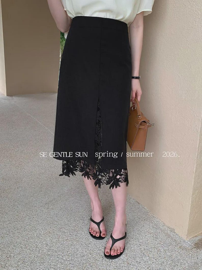 Model wearing a black Tavya high-waist midi skirt with a central front slit, revealing exquisite black floral lace, in a bright, minimalist setting.