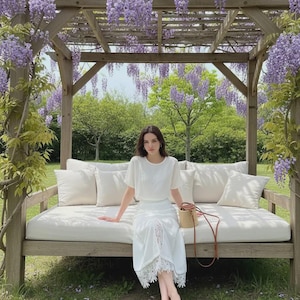 Crisp white high-waist midi skirt with a front slit and romantic floral lace underlay, worn by a model sitting in a wisteria garden.