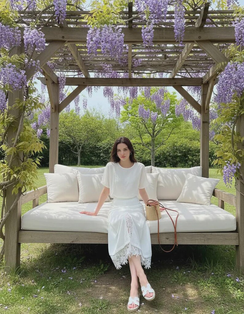 Crisp white high-waist midi skirt with a front slit and romantic floral lace underlay, worn by a model sitting in a wisteria garden.