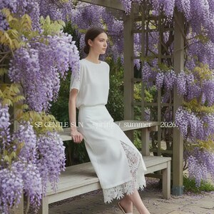 Elegant model wears white Tavya high-waist midi skirt, featuring a front slit revealing intricate floral lace, while sitting on a bench amidst purple wisteria.
