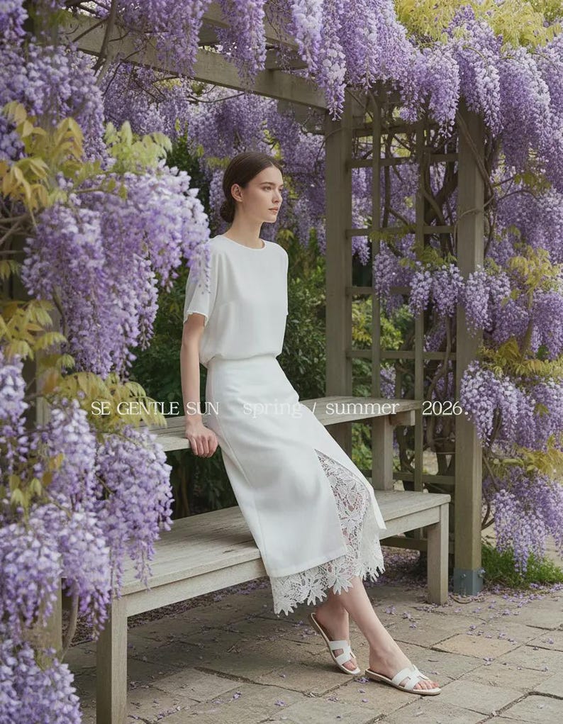 Elegant model wears white Tavya high-waist midi skirt, featuring a front slit revealing intricate floral lace, while sitting on a bench amidst purple wisteria.