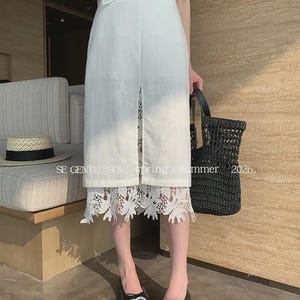 Model wearing a white Tencel blend high-waisted midi skirt with a front slit, revealing an intricate white floral lace underlay.