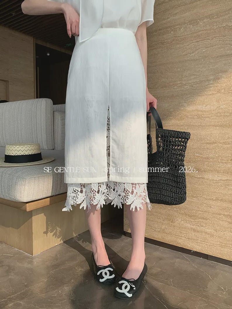 Model wearing a white Tencel blend high-waisted midi skirt with a front slit, revealing an intricate white floral lace underlay.