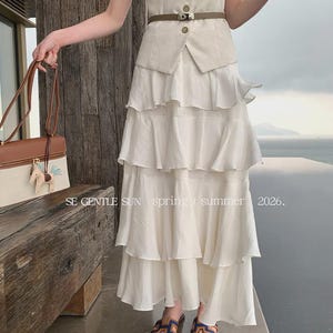A woman wears the Sykas off-white tiered Tencel maxi skirt, featuring five flowing tiers and a crinkled texture, styled outdoors overlooking the sea.