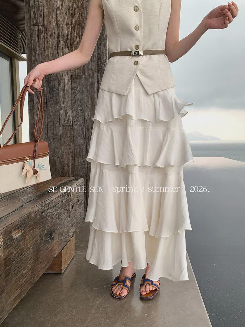 A woman wears the Sykas off-white tiered Tencel maxi skirt, featuring five flowing tiers and a crinkled texture, styled outdoors overlooking the sea.