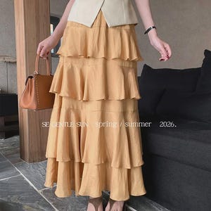 Model wears a Golden Dawn tiered Tencel maxi skirt with a crinkled texture and flowing ruffles, styled with a cream vest, belt, and brown sandals in a refined indoor setting.