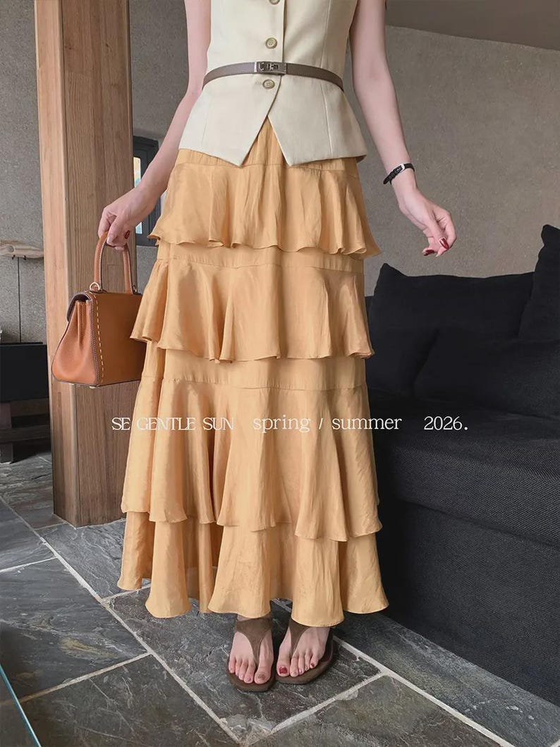 Model wears a Golden Dawn tiered Tencel maxi skirt with a crinkled texture and flowing ruffles, styled with a cream vest, belt, and brown sandals in a refined indoor setting.