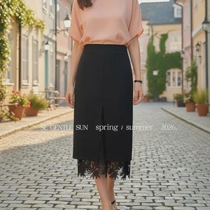 Model in a black high-waisted midi skirt with a front slit showing floral lace detail, walking on a cobblestone street.