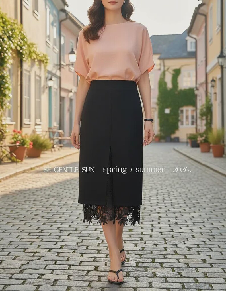 Model in a black high-waisted midi skirt with a front slit showing floral lace detail, walking on a cobblestone street.