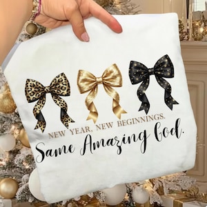 May include: White decorative pillow featuring three bow designs: leopard print, gold, and black with gold stars. The text reads "NEW YEAR, NEW BEGINNINGS. Same Amazing God."