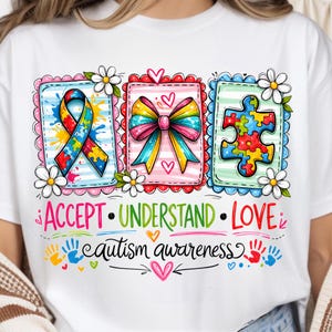 May include: White t-shirt featuring a colorful graphic design with the words "ACCEPT, UNDERSTAND, LOVE" and "autism awareness." The design includes a ribbon, a bow, and a puzzle piece, all in vibrant colors, with hearts and flowers.
