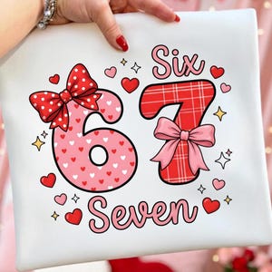 Puede incluir: Cojín cuadrado blanco con los números "6" y "7" en rosa y rojo, decorado con lazos, corazones y estrellas. La palabra "Six" está encima del número 7, y la palabra "Seven" está debajo. El número 6 está cubierto de corazones.