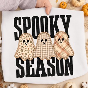 Può includere: Una t-shirt bianca con la scritta nera SPOOKY SEASON e tre illustrazioni di fantasmi. Il fantasma a sinistra ha un motivo floreale, quello centrale un motivo pied-de-poule e quello a destra un motivo a quadri nei toni del marrone e del beige.