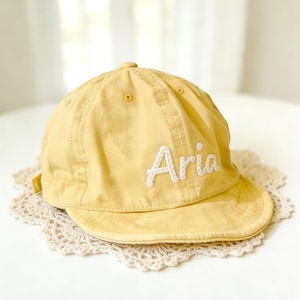 Op de afbeelding: Een lichtgele baseballpet met een gebogen klep. De naam "Aria" is in het wit op de voorkant geborduurd. De pet is gemaakt van een zachte stof en heeft een klein knoopje bovenop. De pet ligt op een wit kanten kleedje.