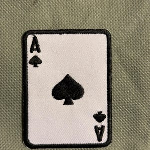 May include: A white Ace of Spades playing card patch with a black border. The card features a large black spade symbol in the center, with smaller spade symbols and the letter "A" at the top and bottom.