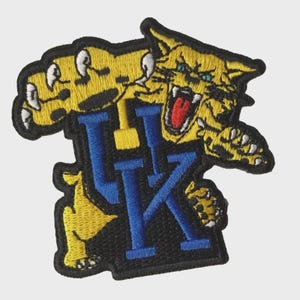 May include: Embroidered patch featuring a snarling yellow wildcat with blue and yellow "UK" lettering on a black background. The wildcat has green eyes and a red mouth, with white claws. The patch is outlined in black.