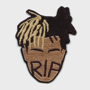 Pode incluir: Patch bordado com um rosto estilizado com olhos fechados e um penteado distinto. O patch tem cores castanhas e bege, com as letras "RIP" bordadas na parte inferior. Ideal para roupas ou acessórios.
