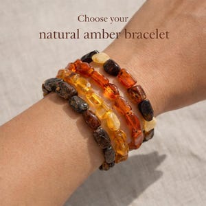 Op de afbeelding: Vier natuurlijke barnsteenarmbanden in verschillende tinten bruin, geel en oranje worden getoond op een pols. De armbanden zijn gemaakt van onregelmatige, gepolijste barnsteen kralen. Boven de armbanden staat de tekst: "Kies je natuurlijke barnsteenarmband."