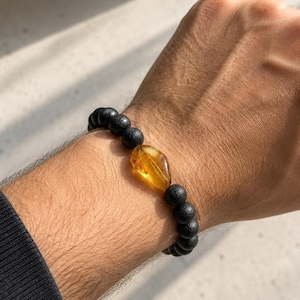 May include: A beaded bracelet featuring a series of matte black spherical beads and a single, central, polished amber-coloured stone with a translucent, irregular, organic shape. The beads are strung together in a continuous circular design.