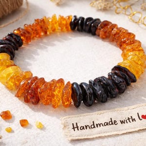 May include: An amber bracelet featuring a mix of dark brown, orange, and yellow beads. The bracelet is displayed on a white surface with a tag that reads "Handmade with love."